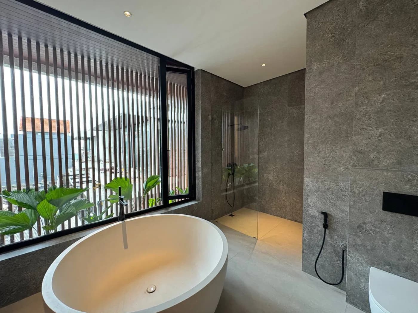 Freestanding bathtub by a timber-slat window with tropical plants